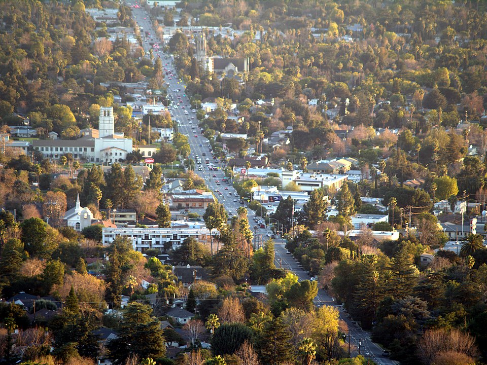 Aerial view of a suburban area with a central road lined by trees and buildings. The setting sun casts a warm glow, creating a tranquil atmosphere.