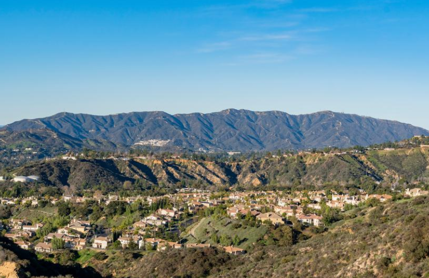 A small residential neighborhood is nestled in a hilly landscape with lush greenery, under a clear blue sky, and backed by distant mountain ranges.